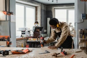 Carpenter using tools in a workshop with a colleague in the background.